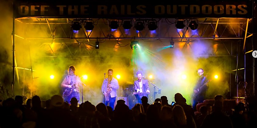 Jeff playing saxophone under stage lights at Off the Rails Outdoors, with the crowd in silhouette